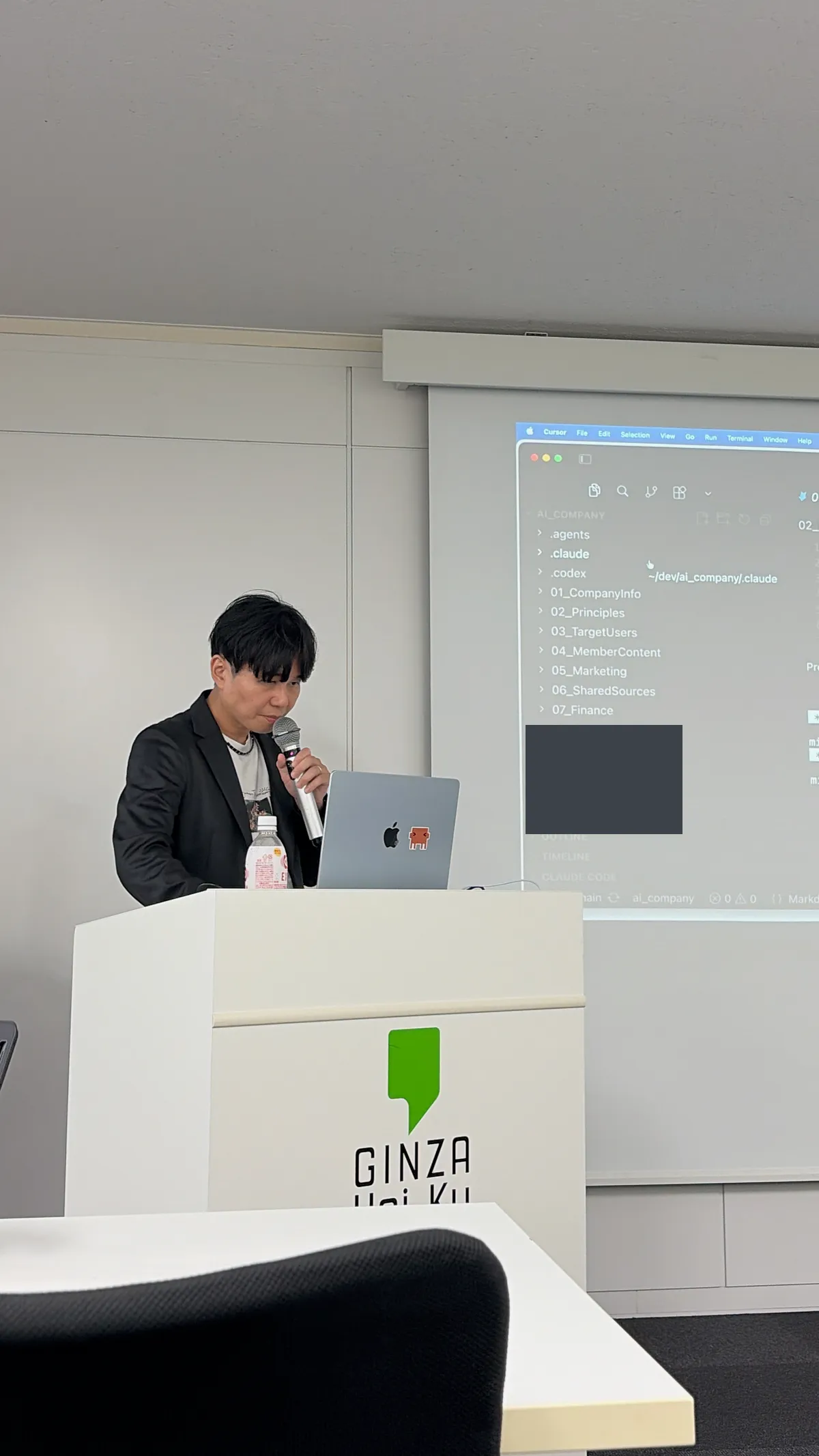 スクリーンにCursorの画面を投影し、自社リポジトリのフォルダ構成を説明するみやっち🧑‍💻。左のサイドバーに .agents / .claude / .codex / 01_CompanyInfo / 02_Principles / 03_TargetUsers / 04_MemberContent / 05_Marketing / 06_SharedSources / 07_Finance などのフォルダが並んでいる