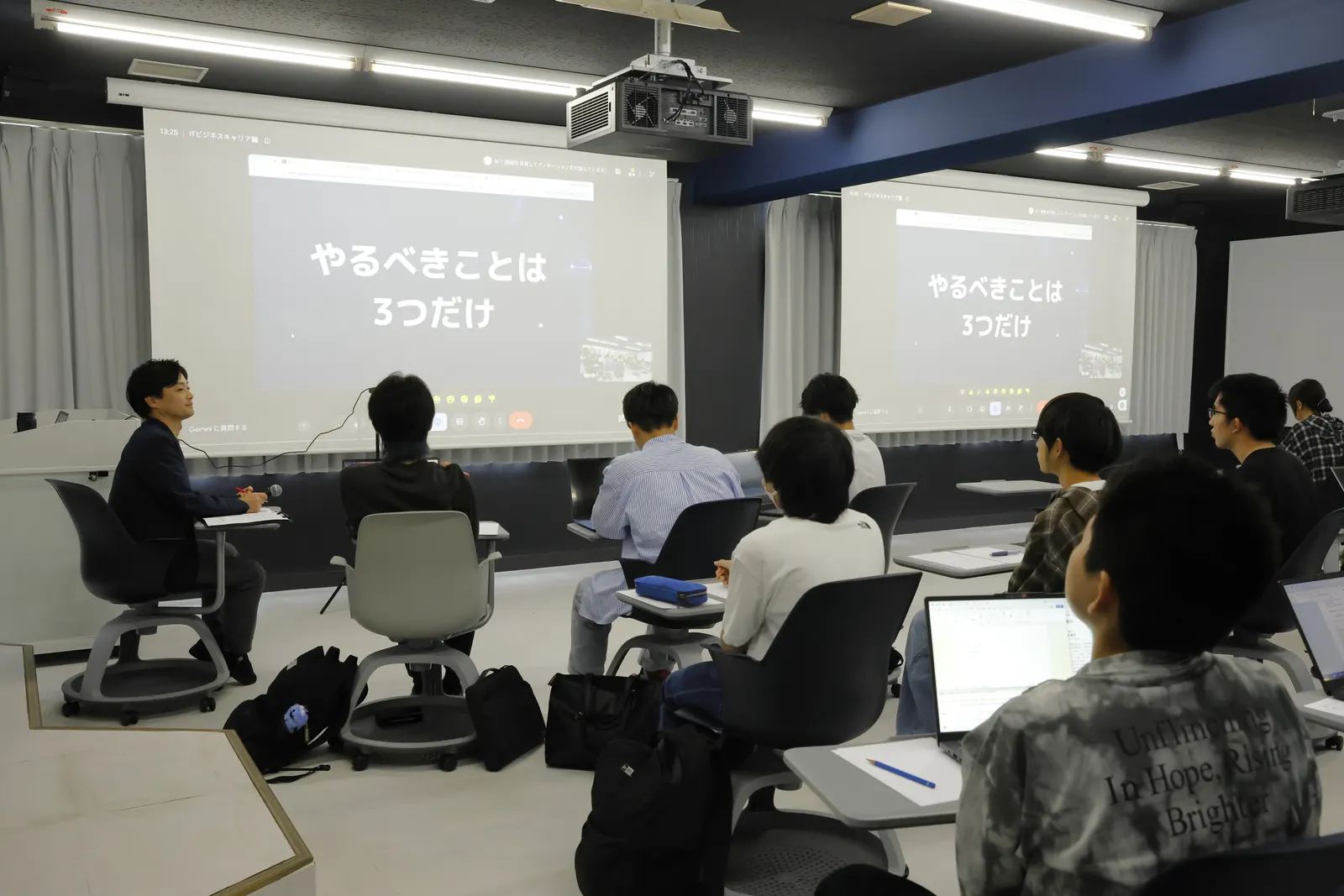 「やるべきことは3つだけ」と映し出された教室の2つのスクリーンと、聴講する学生たち